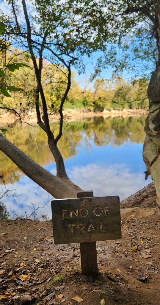 Small sign stating "End of Trail" stuck into dirt trail with body of water directly behind it.