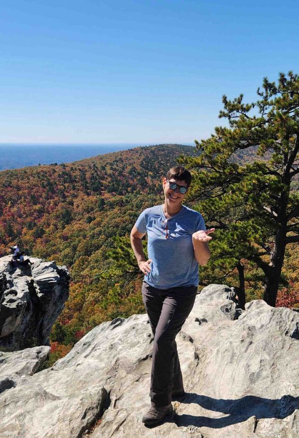 White female in blue t-shirt and gray pants on rock outcropping in front of background of fall foliage.
