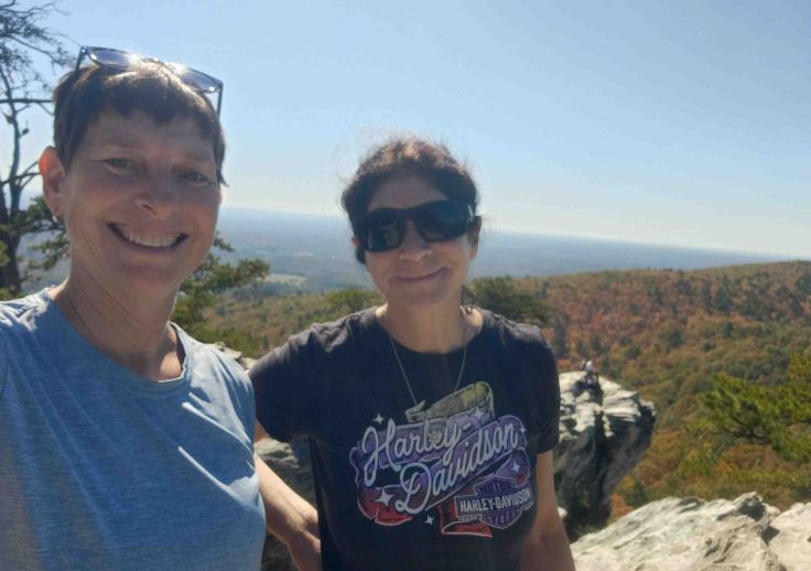 Two white females in short sleeve t-shirts standing on outcropping of rock in front of forest of red, yellow, orange and green foliage.