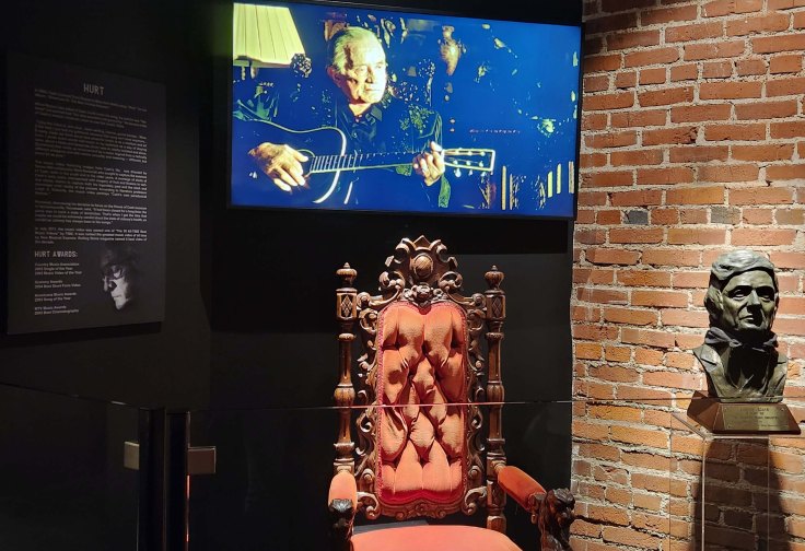 Video of Johnny Cash singing the song ‘Hurt’ over a chair depicting a throne, next to a bust of Johnny Cash and a placard describing the history and significance of the song, written by Trent Reznor.