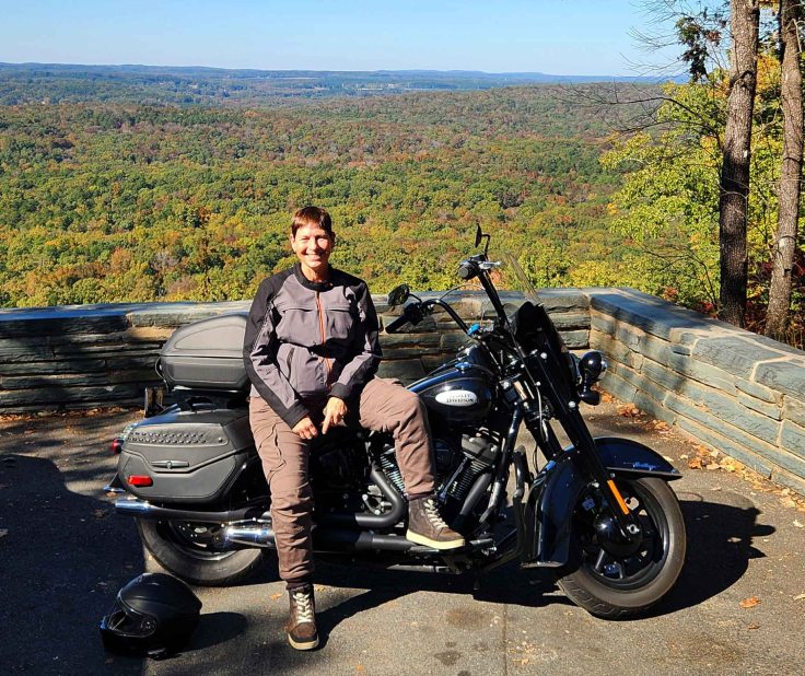 White female in riding gear sitting on Harley motorcycle in foreground with sea of red, orange, yellow and green foliage in background.
