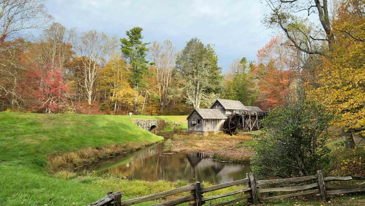 Historic wooden Mabry Mill nestled amongst green, yellow, and red leaved trees, reflected in a pond.