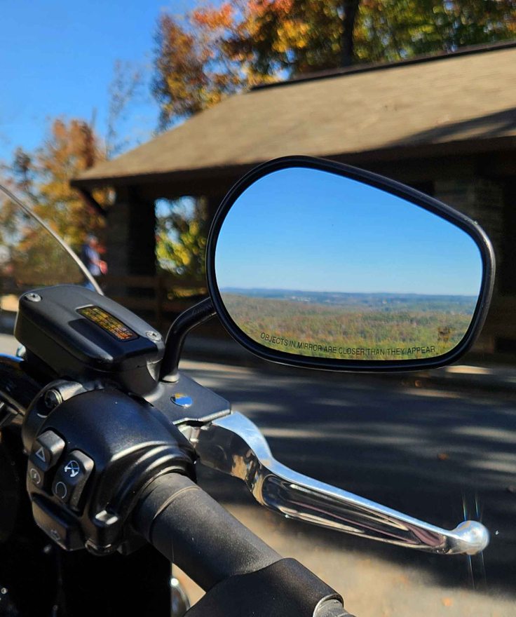 Motorcycle rear view mirror reflecting landscape of fall foliage.