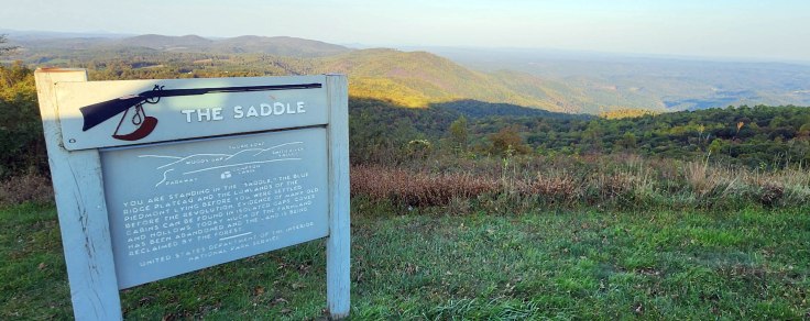 Blue Ridge Parkway sign in the foreground describing The Saddle Overlook with the Virginia Peidmont lowlands in the background